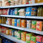 Cesta básica com alimentos como arroz, feijão e vegetais em supermercado brasileiro, ilustrando queda de preços nas capitais.