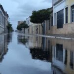 Rua inundada em cidade brasileira durante chuva intensa, com nuvens escuras, representando previsão do Inmet de chuvas acima da média e eventos extremos em janeiro.