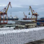 Porto brasileiro com pilhas de sacos de nitrato de amônio, guindastes e navios, refletindo alta nos preços devido à guerra no Oriente Médio.