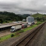 Trilhos de trem em paisagem rural de SC com granjas de frango e silos, representando Ferrovia do Frango e gargalos logísticos no agronegócio.